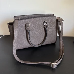 Brun/beige axelväska med blommigt innerfoder - Säljer en stilren brun axelväska med silverfärgade detaljer. Väskan har både handtag och justerbar axelrem. Insidan är fodrad med blommigt tyg och har flera fack. Väskan kan slutas med en dragkedja! 