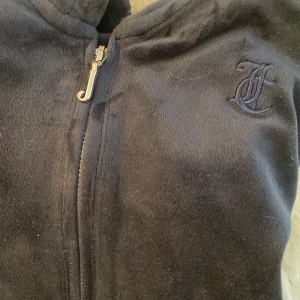 Mörkblå velour hoodie från Juicy Couture och byxor - Mysig mörkblå hoodie i velour från Juicy Couture med dragkedja framtill och broderad JC-logga på bröstet. Jackan har huva, känguruficka och klassisk passform. Perfekt för chill dagar eller när du vill ha en skön och trendig look. Byxorna följer med. Storleken är 14-15 men sitter som en xs-s