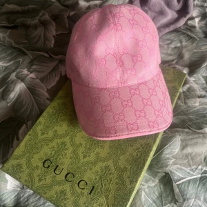 Rosa keps från Gucci i bomull - Säljer en rosa keps från Gucci med ikoniskt GG-mönster över hela kepsen. Kepsen är tillverkad i 100% bomull och har justerbar rem baktill med Gucci-logga. Perfekt accessoar för att lyfta din outfit med lyxig känsla. Jag tar gärna emot prisförslag 