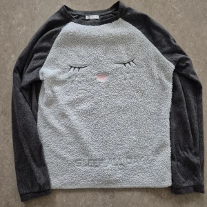 Tröja - Supermjuk och fluffig fleece-tröja från Cubus i grått och vitt med broderat sött ansikte och texten 'SLEEP ALL DAY' framtill. Långärmad och perfekt för chill dagar hemma. Tillverkad i 100% polyester för extra mysfaktor.