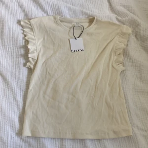 Beige ribbad topp med volangärm Zara - Supersöt beige ribbad topp från Zara med volangdetaljer på axlarna. Toppen har rund halsringning och är ärmlös med snygga volanger som ger en extra touch. Perfekt för dig som gillar en enkel men ändå trendig stil.