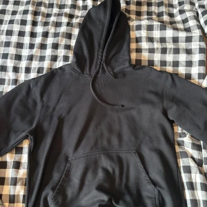 Svart hoodie från Oxide i storlek M - En klassisk svart hoodie från Oxide med huva och känguruficka framtill. Tröjan har långa ärmar och dragsko i huvan. Perfekt för chill dagar eller när du vill ha en enkel och stilren look.