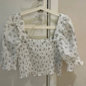 Vit blommig blus med puffärm - Supersöt vit blus med små blommor i lila och grönt. Toppen har puffiga trekvartsärmar, smockad kropp och volangdetaljer längs axelbanden. Knytning i nacken för extra detalj. Perfekt för en romantisk look och passar till både jeans och kjol.