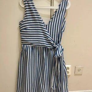 Randig omlottklänning i bomull - Superfin ärmlös kortklänning med blåvita ränder och omlottmodell. Klänningen har v-ringning och knyt i midjan för en snygg passform. Perfekt för varma dagar och ger en fräsch, somrig vibe. Tillverkad i mjuk bomull som känns skön mot huden.
