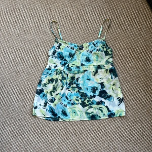 Blommigt linne från Esprit - Supersött linne från Esprit med blommigt mönster i blått, grönt och vitt. Tunt, luftigt tyg med smala reglerbara axelband och knyt framtill. Perfekt för varma dagar när du vill ha en chill och fräsch look. Storlek 34. 