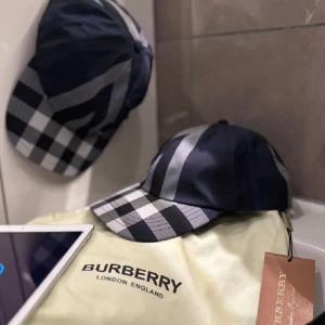 Burberry rutig keps i blått och grått - Snygg keps från Burberry med klassiskt rutigt mönster i blått, grått och vitt. Kepsen har böjd skärm och justerbar passform. Perfekt accessoar för att lyfta din outfit med en lyxig touch. Ikonisk design som känns både modern och tidlös.