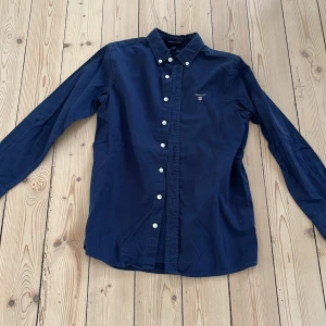 Mörkblå skjorta från Gant, 134/140 - Klassisk mörkblå skjorta från Gant i bomull med button down-krage och vita knappar. Broderad Gant-logga på bröstet ger en clean och stilren look. Perfekt till jeans eller chinos för en snygg vardagsstil.