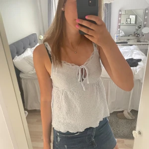 Vit smockad linnetopp med knyt - Supersöt vit linnetopp med smockad struktur och spetskant vid urringningen. Toppen har smala axelband och en söt knytning framtill. Perfekt för varma dagar och ger en romantisk vibe till din outfit.