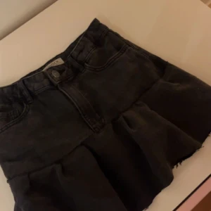 Svart jeanskjol med volang från Ginatricot - Supersnygg svart jeanskjol med volang nertill och rå kant. Klassisk femficksmodell med knapp och dragkedja framtill. Perfekt för dig som gillar en edgy och trendig stil. Den är hemmagjord!