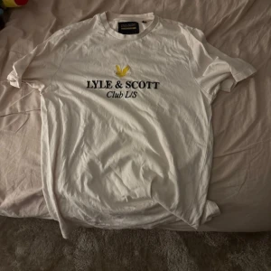 Vit Lyle & Scott t-shirt Club L/S - Vit t-shirt från Lyle & Scott med tryck 'LYLE & SCOTT Club L/S' och den klassiska gula loggan på bröstet. Modellen är relaxed fit och har korta ärmar. Perfekt basic med snygg logga framtill, tillverkad i mjuk bomull.