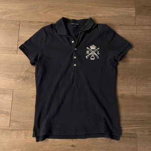 Svart pikétröja från Ralph Lauren - Priset kan diskuteras Klassisk svart pikétröja från Ralph Lauren med broderad RL-logga och detaljer i vitt på bröstet. Tröjan har korta ärmar, krage och knäppning med fyra knappar framtill. Tillverkad i mjuk bomull som känns skön mot huden.