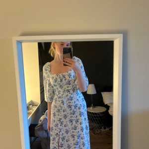 Blommig långklänning med puffärm - Superfin långklänning med blått blommönster på vit botten. Klänningen har puffiga trekvartsärmar, fyrkantig ringning med snörning och en slits framtill. Perfekt för sommarens alla tillfällen!