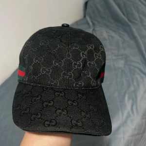 Svart Gucci keps med GG-mönster - Snygg svart keps från Gucci med klassiskt GG-monogram över hela kepsen. Sidorna har gröna och röda ränder som ger den ikoniska Gucci-looken. Justerbar rem bak i svart skinn. Perfekt accessoar för att lyfta din outfit. För mer bilder, frågor eller prisdiskkutering kontakta mig :) Priset kan diskuteras 