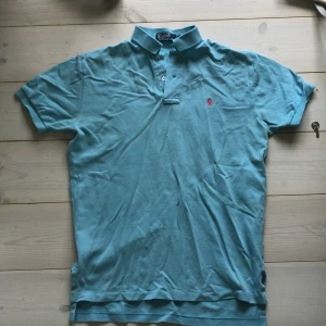 Turkos pikétröja från Ralph Lauren - Snygg turkos pikétröja från Polo Ralph Lauren med klassisk krage och knäppning framtill. Tröjan har korta ärmar och den ikoniska rosa broderade loggan på bröstet. Perfekt för en stilren och avslappnad look.