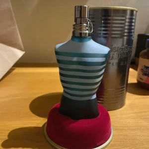 Jean Paul Gaultier Le Male parfym - Ikonisk parfymflaska i form av en manlig torso med blå och turkosa ränder, metallisk spraykork och tillhörande silvrig plåtburk. Flaskan är tillverkad i frostat glas och har en modern, maritim känsla. Perfekt för dig som gillar unika och snygga flaskor.