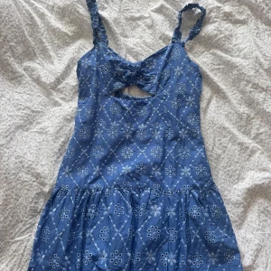 Blå kortklänning med broderi - Supersöt blå kortklänning med vita broderade blommor och smock i ryggen. Klänningen har axelband, en liten cut-out framtill och volang i nederdelen! Aldrig använd!