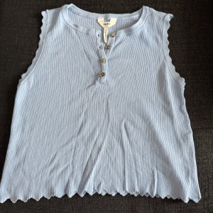 Ljusblå ribbad linnetopp  - Supersöt ljusblå ribbad linnetopp med dekorativa knappar framtill och vågiga kanter vid ärmhål och nederkant. Toppen är ärmlös och har en mjuk, stretchig känsla som gör den riktigt bekväm och snygg till jeans eller kjol.