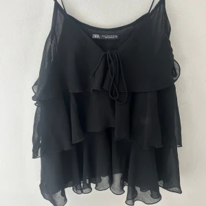 Svart volangig linnetopp från Zara - Supersöt svart linnetopp från Zara med tunna axelband och flera lager volanger. Toppen har v-ringning och knyt framtill för extra detalj. Tunt, flowy tyg som ger en luftig känsla och snyggt fall. Passar S - pris kan absolut diskuteras, säljs för att tröjan inte kommer till användning. 