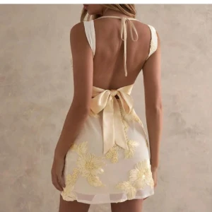 Champagnefärgad blommig kortklänning - Superfin kort klänning i champagnefärg med stora broderade blommor i gult och vitt. Klänningen har öppen rygg, breda axelband och en stor rosett i ryggen. Materialet är tunt och lätt, perfekt för sommarens alla tillfällen.