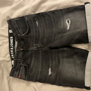 Svarta jeansshorts Jack & Jones - Svarta jeansshorts från Jack & Jones med slitna detaljer och uppvikta benslut. Klassisk femficksmodell med regular fit och snygg tvättad look. Perfekta för dig som gillar en avslappnad och trendig stil.