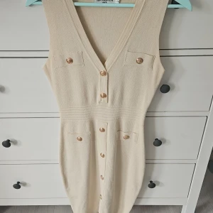 Beige stickad klänning J&H Fashion - Supersnygg beige stickad klänning från J&H Fashion med guldiga knappar framtill, v-ringning och fejkfickor. Ärmlös och figurnära modell som ger en clean och chic look. Perfekt för dig som gillar stilrena detaljer och vill ha något bekvämt men ändå classy.