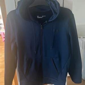 Mörkblå hoodie från Under Armour - Säljer en mörkblå hoodie från Under Armour med dragkedja framtill och huva med snörning. Tröjan har två stora fickor och är perfekt för träning eller chill. Klassisk och stilren design med diskret logga på bröstet.