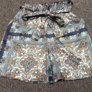 Mönstrade shorts från Gina Tricot - Snygga shorts från Gina Tricot med hög midja och knytband. De har ett unikt paisleyinspirerat mönster i blått, vitt, orange och grått. Luftigt material, perfekt för varma dagar. Styla med en enkel topp för en cool look.