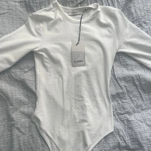 Vit långärmad body från Avora - Supersnygg vit body från Avora med trekvarts ärm. Tight passform som sitter perfekt och ger en clean look. Tillverkad i mjuk bomullsmix och har diskret broderad logga framtill. Perfekt att styla med jeans eller kjol för en enkel men fräsch outfit.