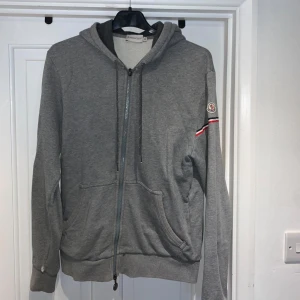 Grå Moncler hoodie med dragkedja - Snygg grå zip up hoodie från moncler. Defects på båda cuffs som man ser i bild 4. Köpte för 5500kr! VIKTIGT säger medium men passar som S