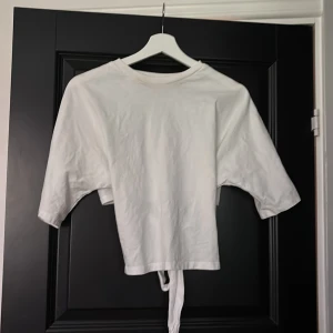Vit croppad topp med knyt i ryggen - Säljer en vit croppad topp i bomull med korta ärmar och snygg knytning i ryggen. Toppen har en enkel och clean look framtill och cool öppen rygg med breda knytband som ger en trendig vibe. Perfekt till jeans eller kjol för en chill stil.