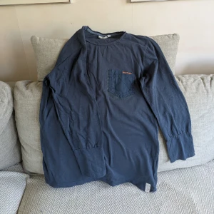 Mörkblå långärmad tröja från Denimwear - Mörkblå ganska tunn långärmad tröja från Denimwear. Tröjan har en rund halsringning och en bröstficka med orange broderad logga. Jag är ca 170 cm för referens. Vid köp av flera klädesplagg av mig samfraktar jag såklart!
