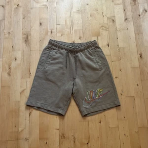 Beige Nike shorts med färgglad logga - Snygga beige shorts från Nike i mjuk bomull med elastisk midja och snörning. Färgglad Nike-logga framtill, två sidofickor och en bakficka. Perfekta för chill dagar eller träning. Klassisk loose fit och skön känsla. Passar folk mellan 163-175
