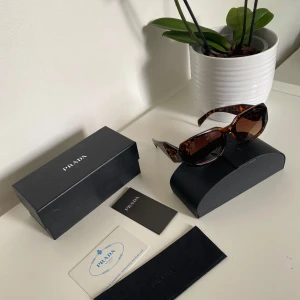 Prada solglasögon brun leopardmönstrad - Snygga solglasögon från Prada med fyrkantig form och brun leopardmönstrad båge. Glasen är bruna och bågarna har guldfärgad Prada-logga på sidan. Kommer med originalfodral, ask och certifikat. Perfekt accessoar för att lyfta din stil.