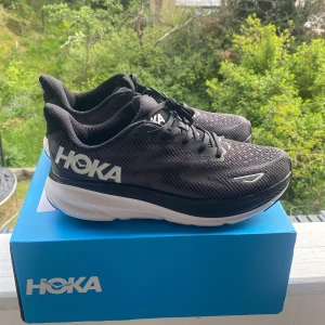 Svarta Hoka Clifton 9 sneakers - Snygga Hoka Clifton 9 sneakers i svart med vita detaljer och stor logga på sidan. Ovandel i mesh för bra andningsförmåga och tjock, dämpad sula för maxad komfort. Perfekta för löpning eller powerwalks. Klassisk snörning och sportig vibe.