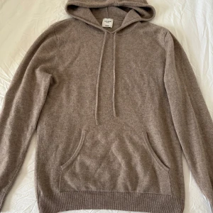 Beige kashmirhoodie från John Henric - Mjuk och lyxig beige hoodie i 100% kashmir från John Henric. Klassisk modell med huva, snörning och känguruficka framtill. Perfekt för dig som gillar stilren och avslappnad look med premiumkänsla.