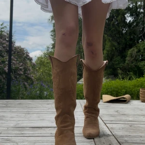 Bruna mocka cowboy boots med klack - Snygga bruna cowboy boots i mocka med klassiska broderade detaljer och spetsig tå. Bootsens höga skaft och blockklack ger en cool western-vibe. Perfekta till både jeans och klänning för en trendig look. Knappt använda, säljer då de är för stora för mig (har vanligast 38-39). Pris kan diskuteras