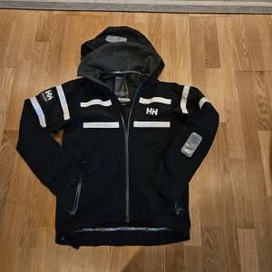 Helly hansen regn och vindjacka  - Mörkblå vindjacka från Helly Hansen med huva, hel dragkedja och vita reflexdetaljer på ärmar, bröst och rygg. Jackan har fyra dragkedjeförsedda fickor fram och två inne fickor, kom gjräna ett bud🙂