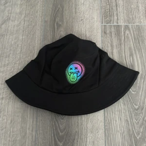 Bucket hat merch Louis Tomlinson - Svart bucket hat merch från Louis Tomlinson med ett färgglatt neon smiley-tryck framtill. Hatten har bred brätte och är tillverkad i mjuk bomull, perfekt för en avslappnad streetstyle. Andvänd ett fåtal gånger.