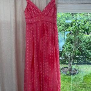 Rosa långklänning med mönster - Söt rosa långklänning med vita geometriska mönster och tunna axelband. Klänningen har omlott-v-ringning och fina spetsdetaljer vid midjan. Tyget är lätt och luftigt, perfekt för varma dagar. Passformen är lös och flowy för en avslappnad vibe.