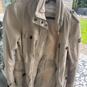 Beige jacka från Peak Performance S - Snygg beige jacka från Peak Performance med flera fickor, axelklaffar och dragkedja framtill. Jackan har en rak passform och är tillverkad i bomull, perfekt för vår och höst. Stilsäker detalj med logotyp på ärmen och knappar på fickorna.