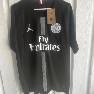 PSG Neymar Jr svart matchtröja - Snygg svart PSG fotbollströja med Neymar Jr och nummer 10 på ryggen. Tröjan har korta ärmar, vit logga och sponsortryck på bröstet samt en diskret rand i mitten. Tillverkad i lätt och ventilerande material, perfekt för fotbollsplanen eller att bära som supporter.