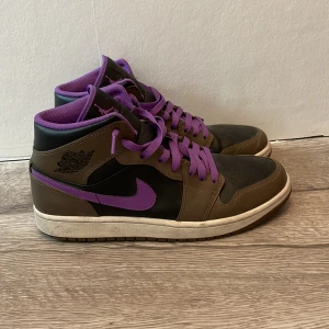 Nike Air Jordan 1 Mid brun/lila - Säljer ett par Nike Air Jordan 1 Mid i brun och svart med lila detaljer och snörning. Skorna har lila swoosh, lila foder och lila skosnören. Klassisk rund tå och platt sula. Perfekta för dig som vill sticka ut med färg och stil. Dom är i ny skick använd typ 2 gånger. Skriv vid funderingar.