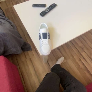 Valentino Garavani vita sneakers - Snygga vita Valentino Garavani sneakers med bred marinblå rem och nitar på hälen. Skorna har rund tå, platt sula och är gjorda i skinn. Perfekta för dig som vill ha en clean och lyxig look med en twist.