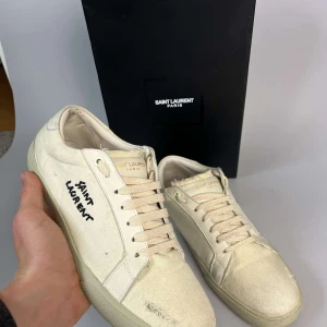 Saint Laurent beiga sneakers i canvas - Snygga beiga sneakers från Saint Laurent i canvas med handskriven logga på sidan. Skorna har rund tå, platt sula och vita snören. Stilsäkra och minimalistiska med diskreta detaljer i skinn och broderad text. Perfekta för dig som gillar exklusiv streetstyle.