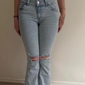 Ljusblå bootcut jeans med slitna knän - Säljer ett par ljusblå bootcut jeans med slitna hål över båda knäna och snygga rosa broderade detaljer på bakfickorna. Jeansen är lågmidjade. Köpta för 699, men säljs för 460 men pris kan diskuteras.