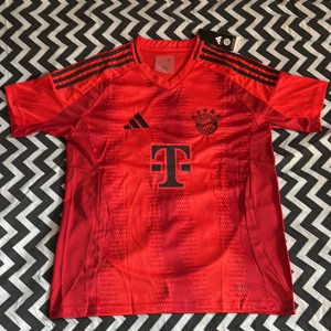 Röd FC Bayern München tröja Adidas M - Snygg röd FC Bayern München fotbollströja från Adidas med svarta detaljer och klubbmärke på bröstet. Tröjan har korta ärmar, V-ringning och klassiska Adidas-ränder på axlarna. Tillverkad i lätt och ventilerande material, perfekt för match eller träning.