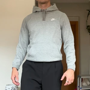 Grå Nike hoodie - Snygg grå hoodie från Nike med klassisk logga på bröstet. Tröjan har huva med snörning och en stor magficka. Tillverkad i mjuk bomullsmix, perfekt för chill eller träning. Långärmad och normal passform.