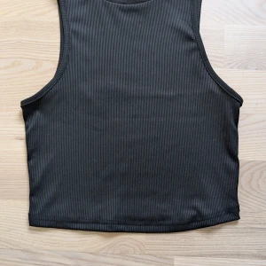 Svart ribbad croppad linnetopp - Snygg svart croppad linnetopp med ribbad struktur. Toppen är ärmlös och har en enkel, stilren design som passar perfekt till jeans eller kjol. Perfekt för dig som gillar en clean och modern look. Använd en gång.