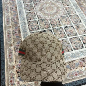 GG keps i färgen beige snygg nytt - Snygg keps från i beige och brunt med klassiskt GG-monogram över hela kepsen. På sidan finns den ikoniska gröna och röda randen. Kepsen är tillverkad i bomull och har justerbar rem baktill. Perfekt för dig som vill sticka ut med en lyxig accessoar.