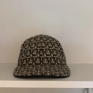 Ferragamo keps  - Snygg keps från Salvatore Ferragamo i beige och svart med ikoniskt mönster över hela kepsen. Justerbar rem baktill med metallspänne. Tillverkad i Italien och i bomullsmaterial. Perfekt accessoar för att lyfta din outfit.